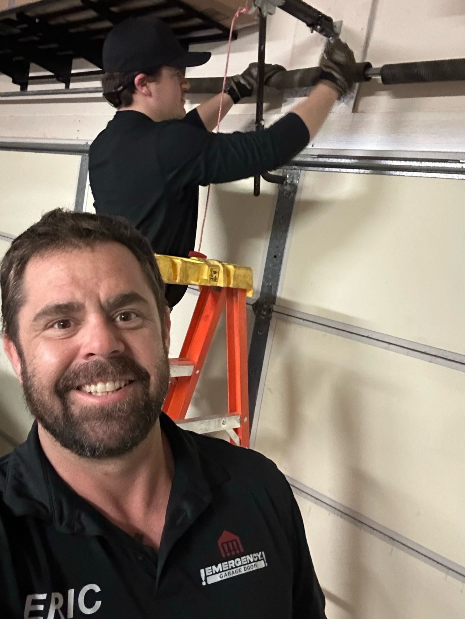 A man with a beard is standing next to a man on a ladder fixing a garage door.