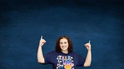 A woman in a blue t-shirt is standing in front of a blue background.