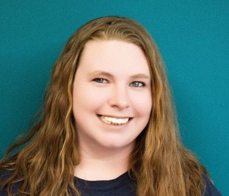 A woman with long brown hair is smiling and wearing a blue shirt.