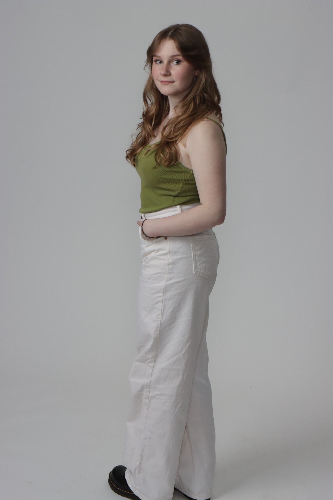 A young woman in a green tank top and white pants is standing in front of a white background.