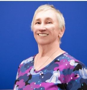 A woman in a purple scrub top is smiling in front of a blue background.