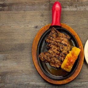 A skillet filled with steak and corn on the cob on a wooden table.
