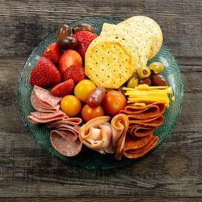 A glass plate filled with meat , cheese , crackers , strawberries , olives and tomatoes.