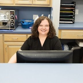 A woman is smiling while sitting at a desk in front of a computer monitor