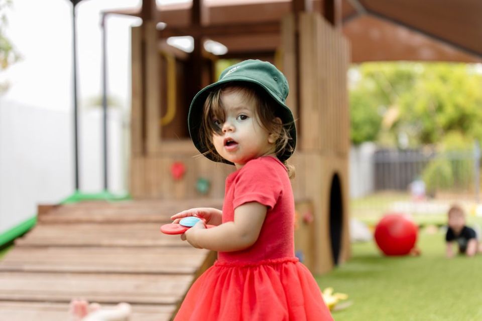 A little girl in a red dress and green hat is standing in front of a playground at Discover Early Learning.