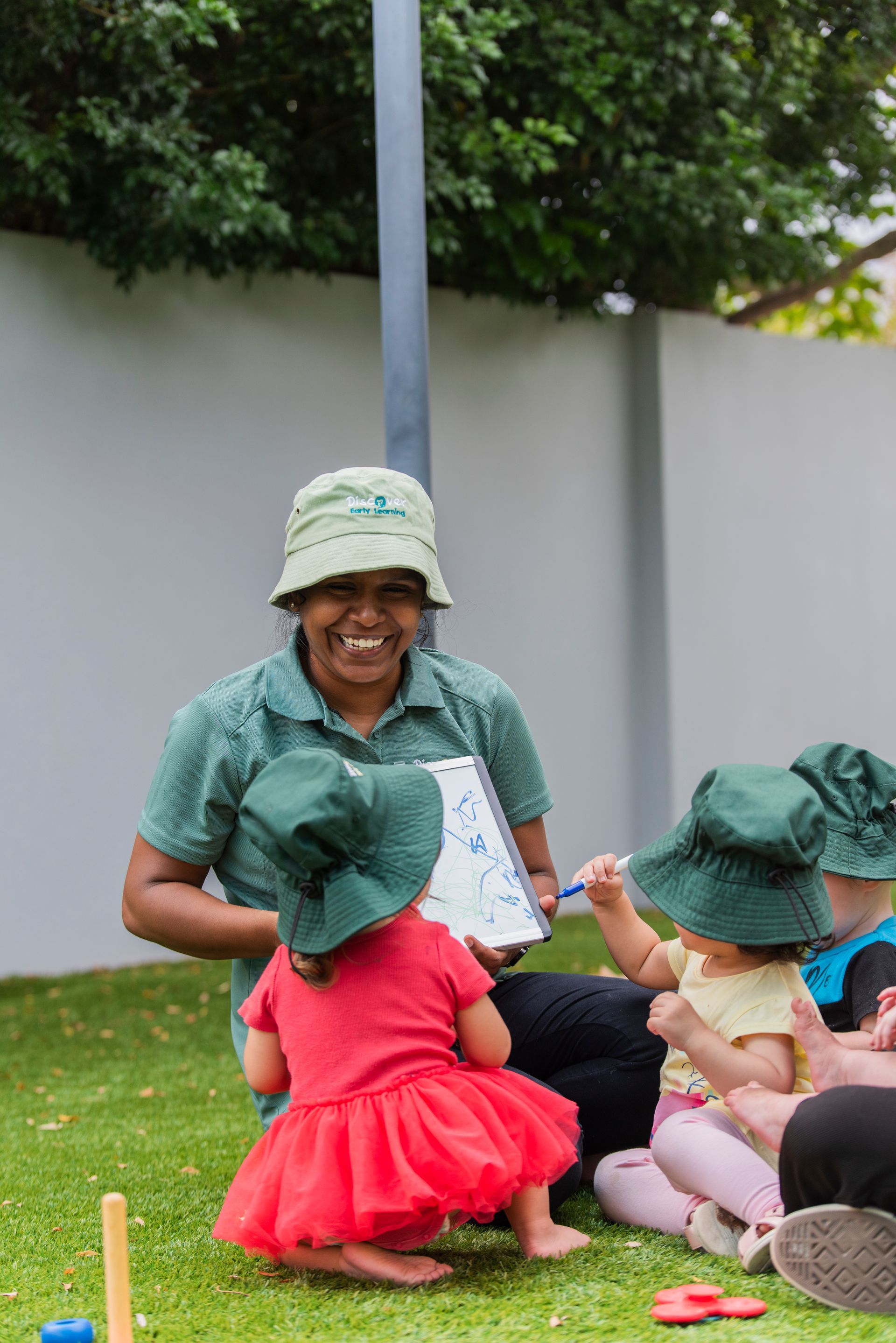A woman is sitting on the grass, reading a book to a group of children at Discover Early Learning Osborne Park Centre.