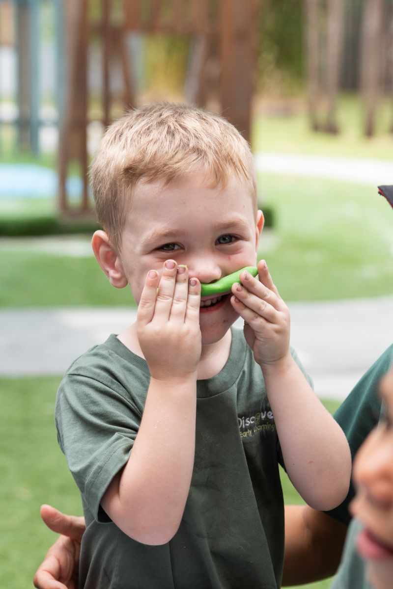 A young boy is biting into a green object at Discover Early Learning.