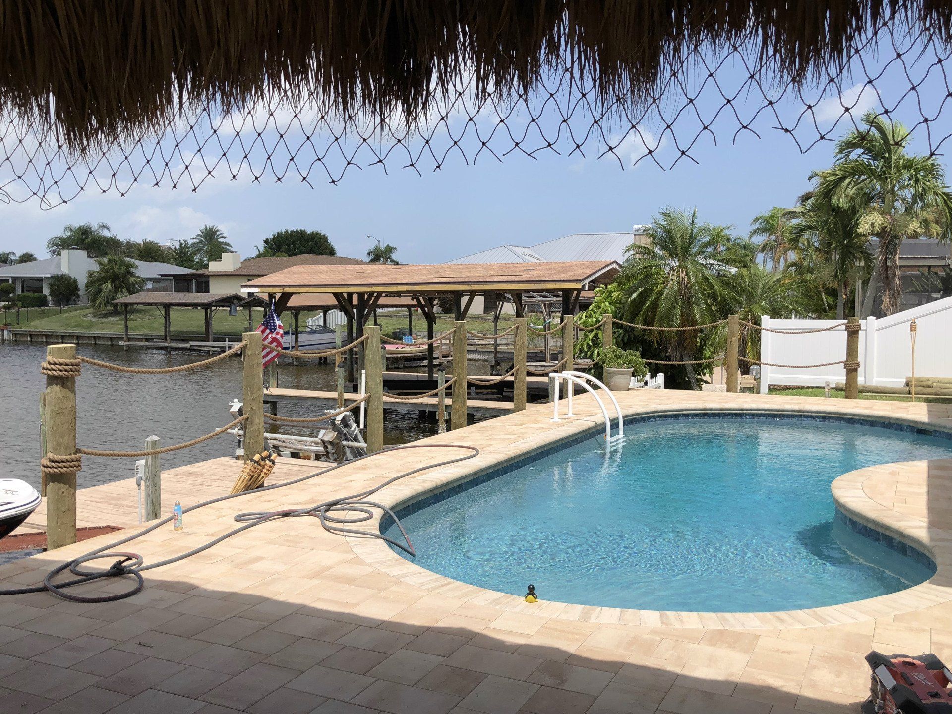 Poolside view with dock, canal, and boat, under a thatch roof. Blue water, sunny day.
