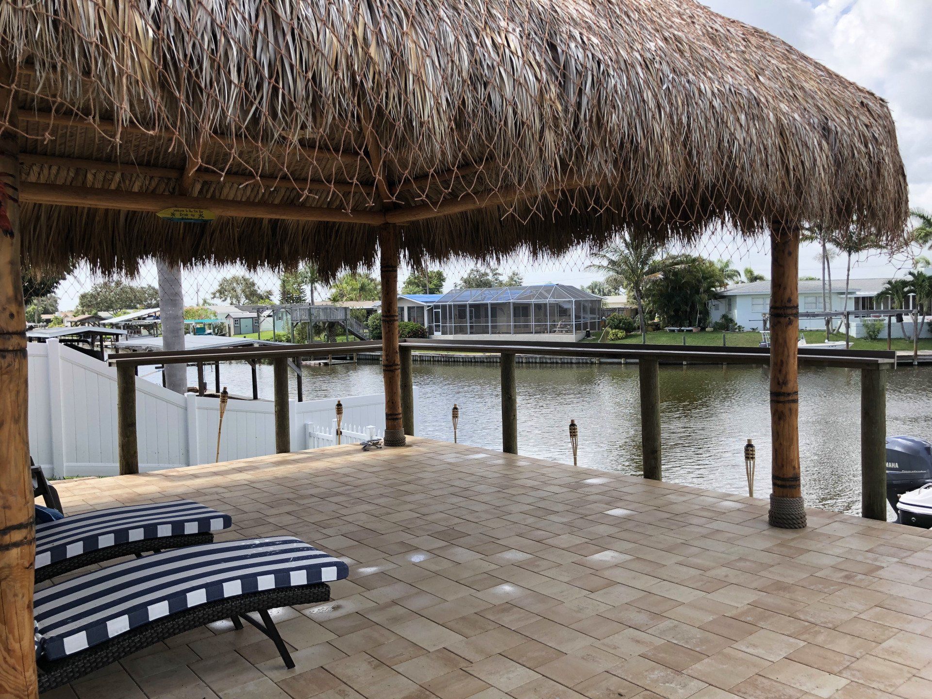 Tiled patio with a thatched roof, two striped lounge chairs, overlooking a canal with houses in the background.
