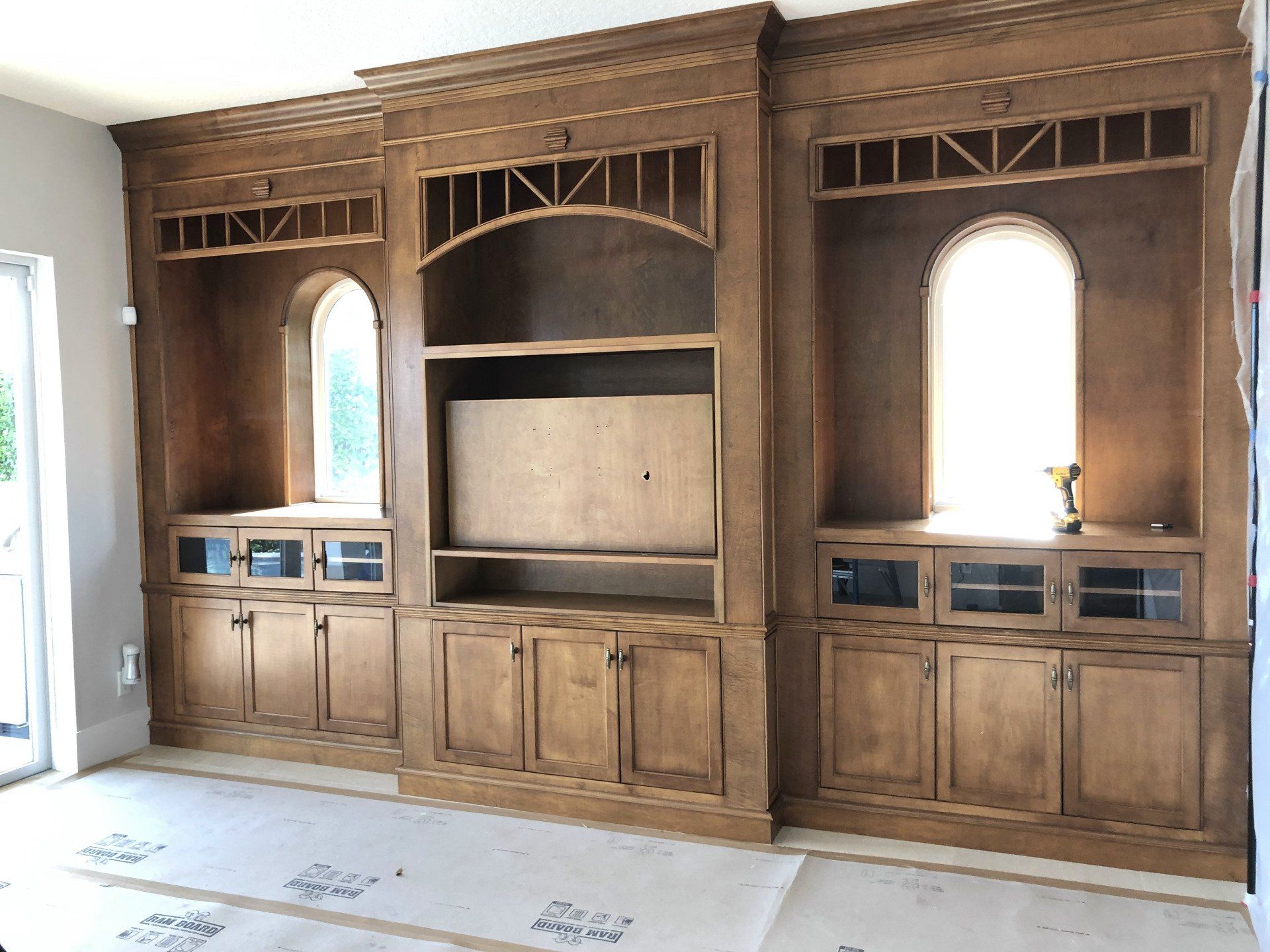Wooden built-in shelving unit with arched windows and storage cabinets, installed in a room with unfinished flooring.