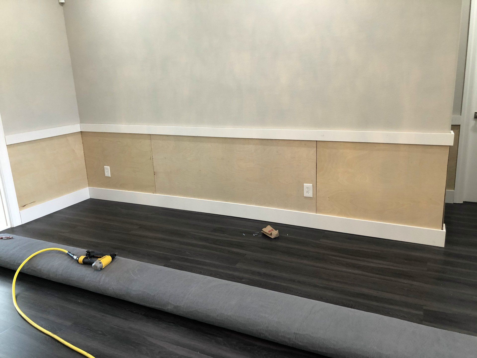 Room with installed dark wood-look flooring and wainscoting on walls, with a roll of gray carpet and tools.
