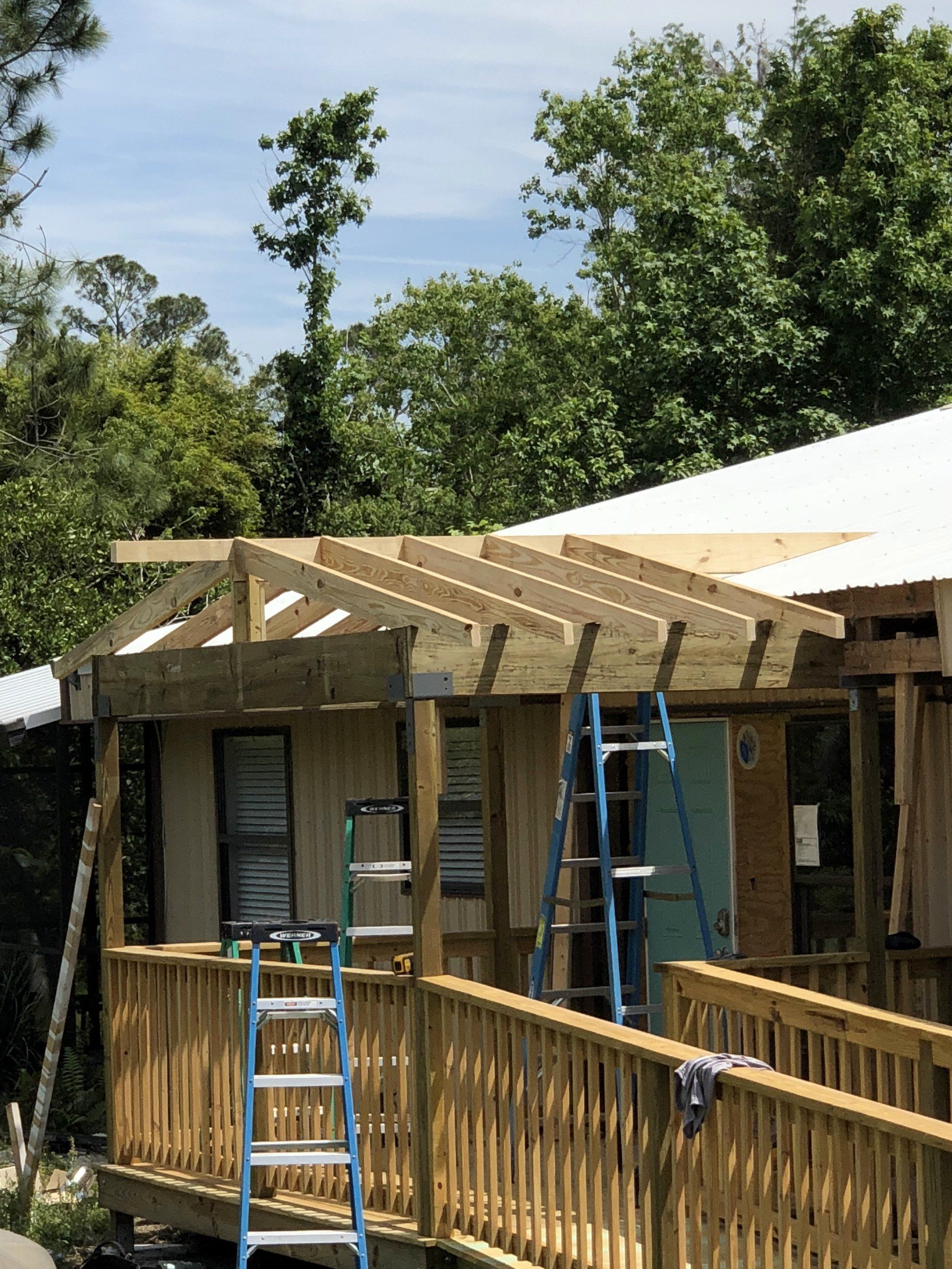 Construction of a wooden porch with a ramp and roof, blue ladder in the foreground, trees in the background.