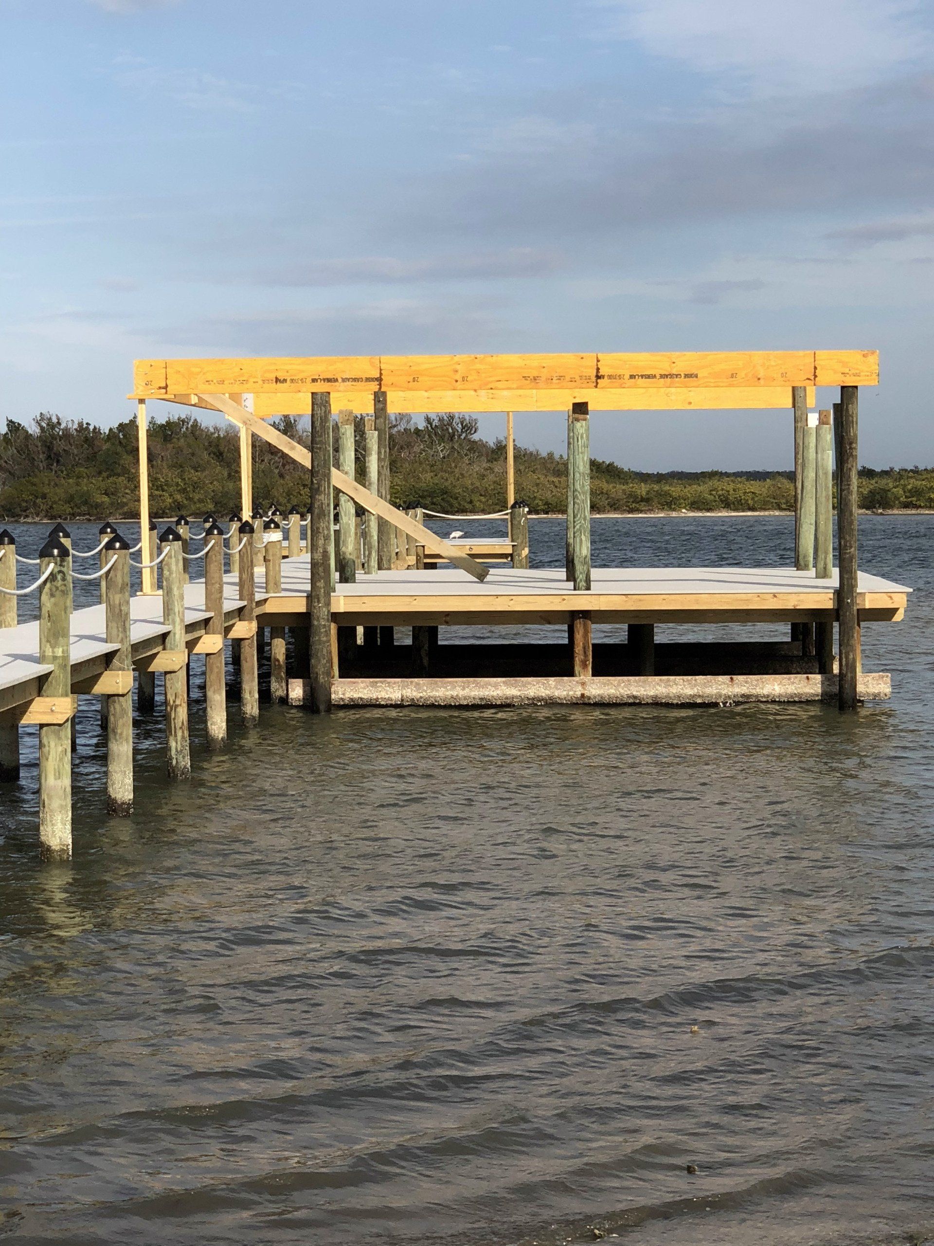 Wooden dock extending into water with a covered platform; light blue sky and coastal vegetation in the background.
