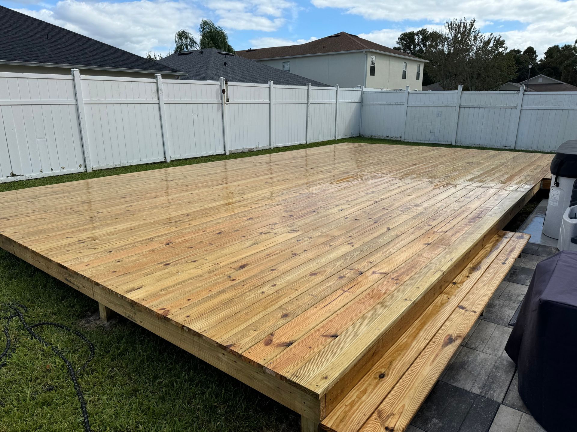 Wooden deck with steps, wet from cleaning, in a backyard with white fence.
