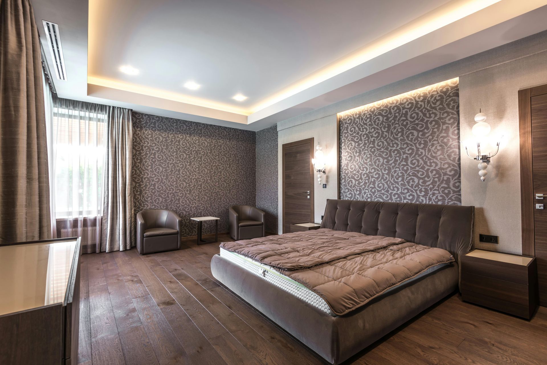 Luxurious bedroom with king-sized bed, dark wood floor, patterned wall, chairs, and sconces.