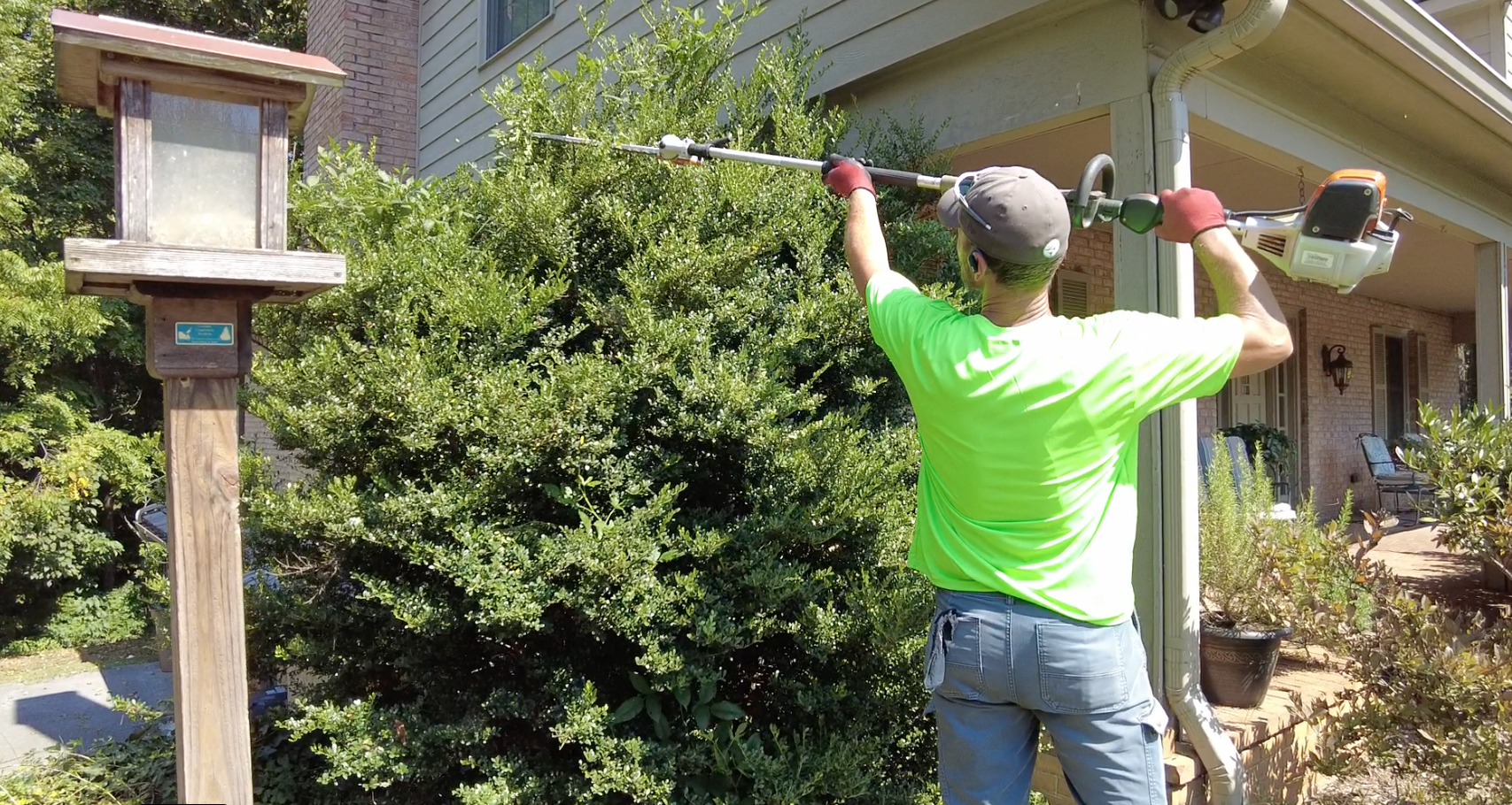 Man trimming bushes with a string trimmer near a birdhouse by a house.