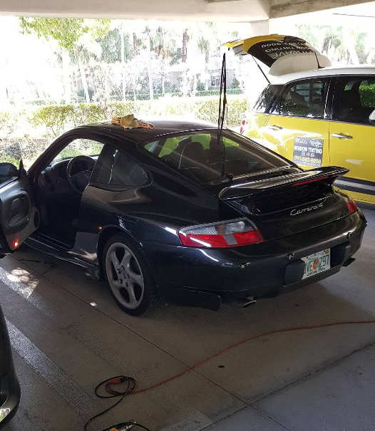 Black Porsche coupe with door open, parked under a shelter next to a yellow SUV with its trunk open.