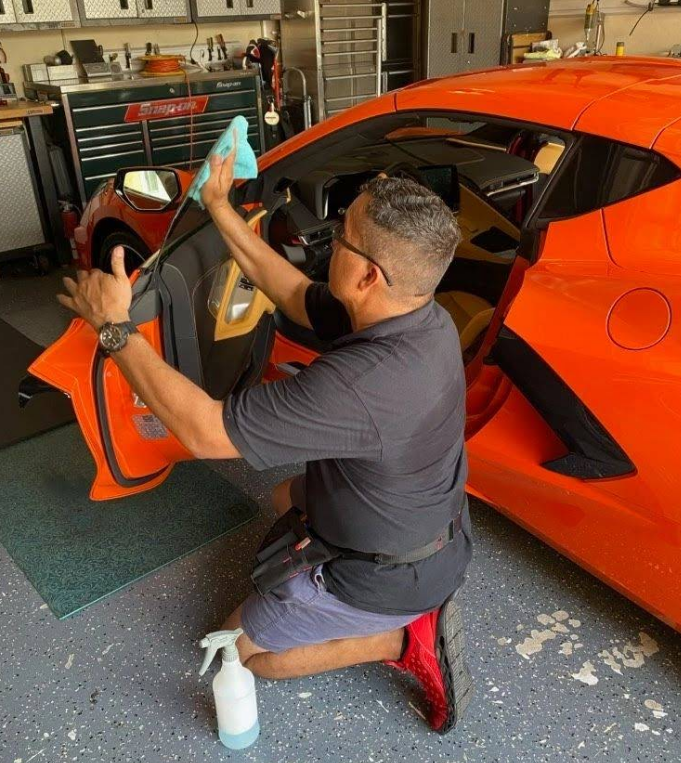 Man cleaning the interior of an orange sports car, kneeling with spray bottle, in a garage.