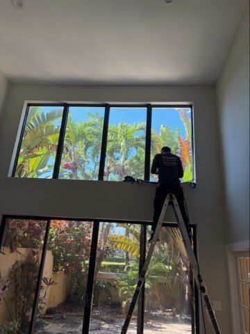 Man on ladder installing window tint on a large window overlooking a yard.