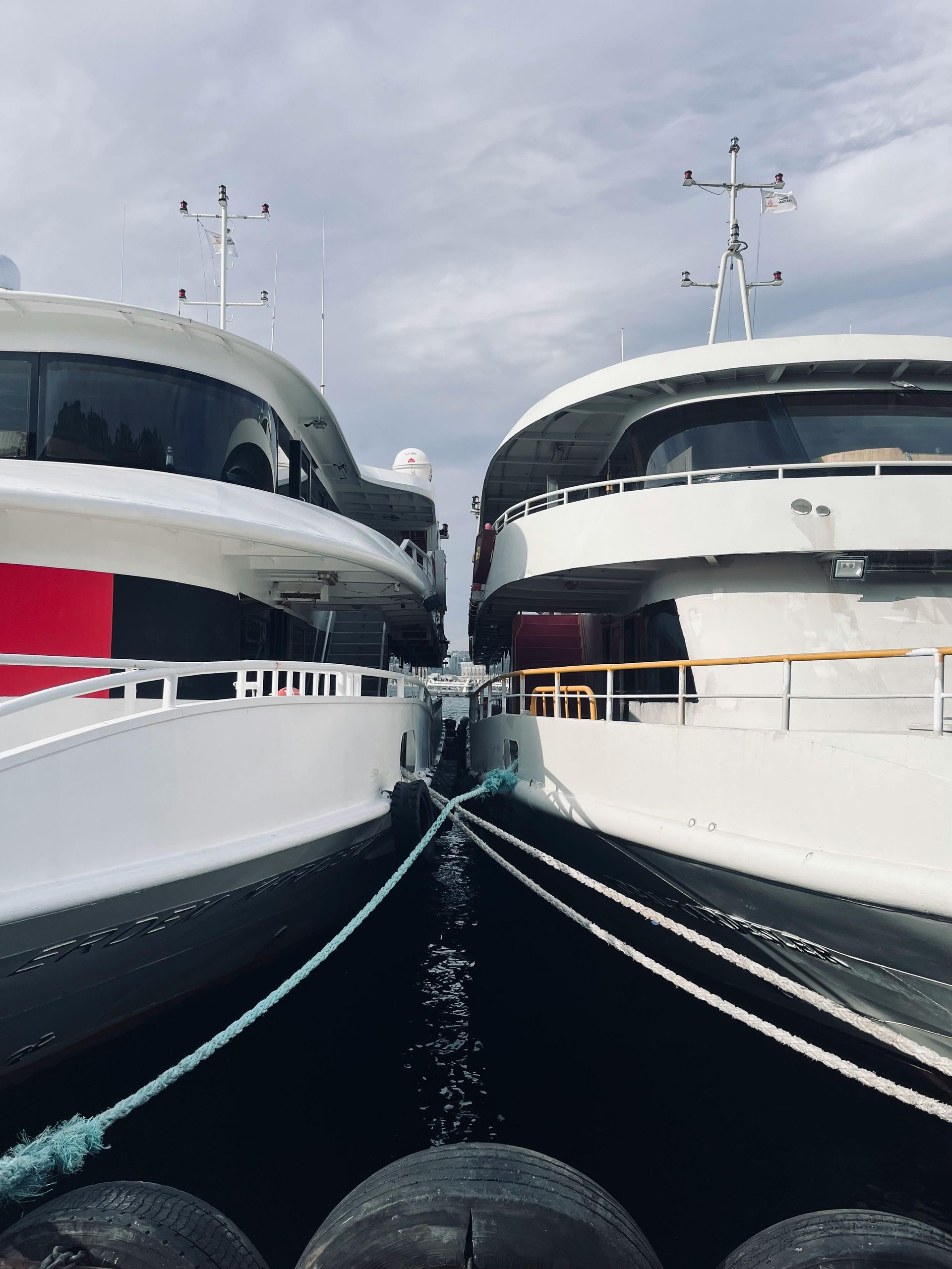 Two large white yachts are docked side-by-side, sharing a narrow space. Blue rope runs between them.