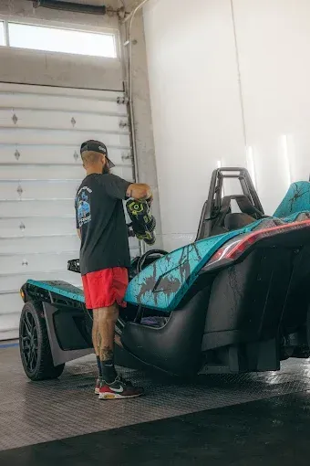 Person cleaning a teal and black Slingshot three-wheeled vehicle in a car wash.