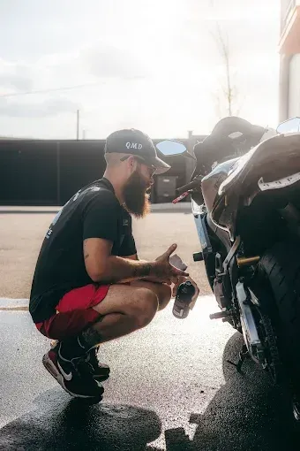 Person squats, cleaning a black motorcycle with a spray bottle, outdoors on a sunny day.