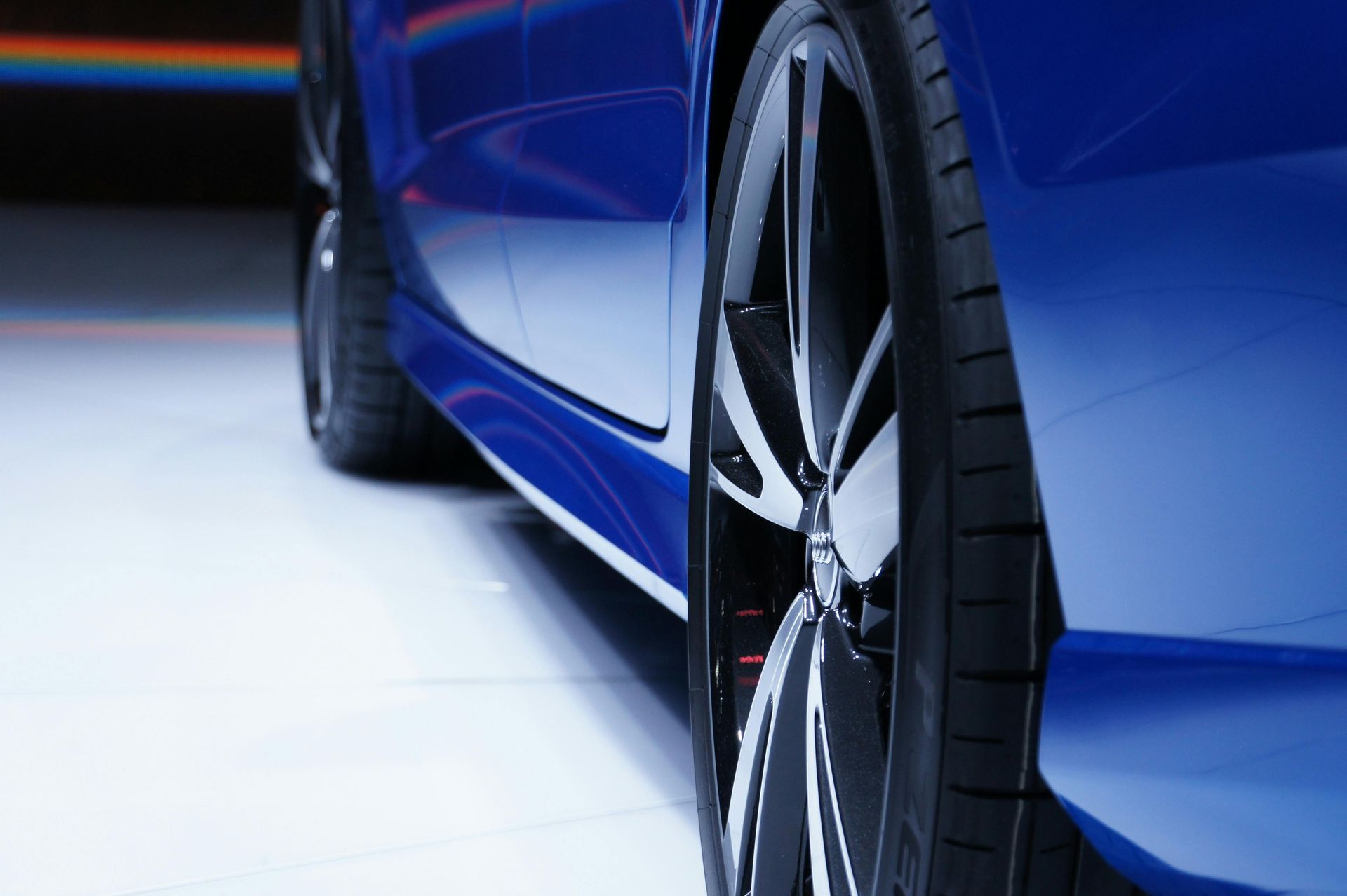 Blue car's side, focus on tire and rim. Shiny, modern design. White floor, dark background.