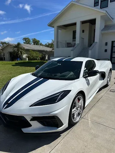 White Corvette with dark racing stripes parked in front of a modern white house on a sunny day.