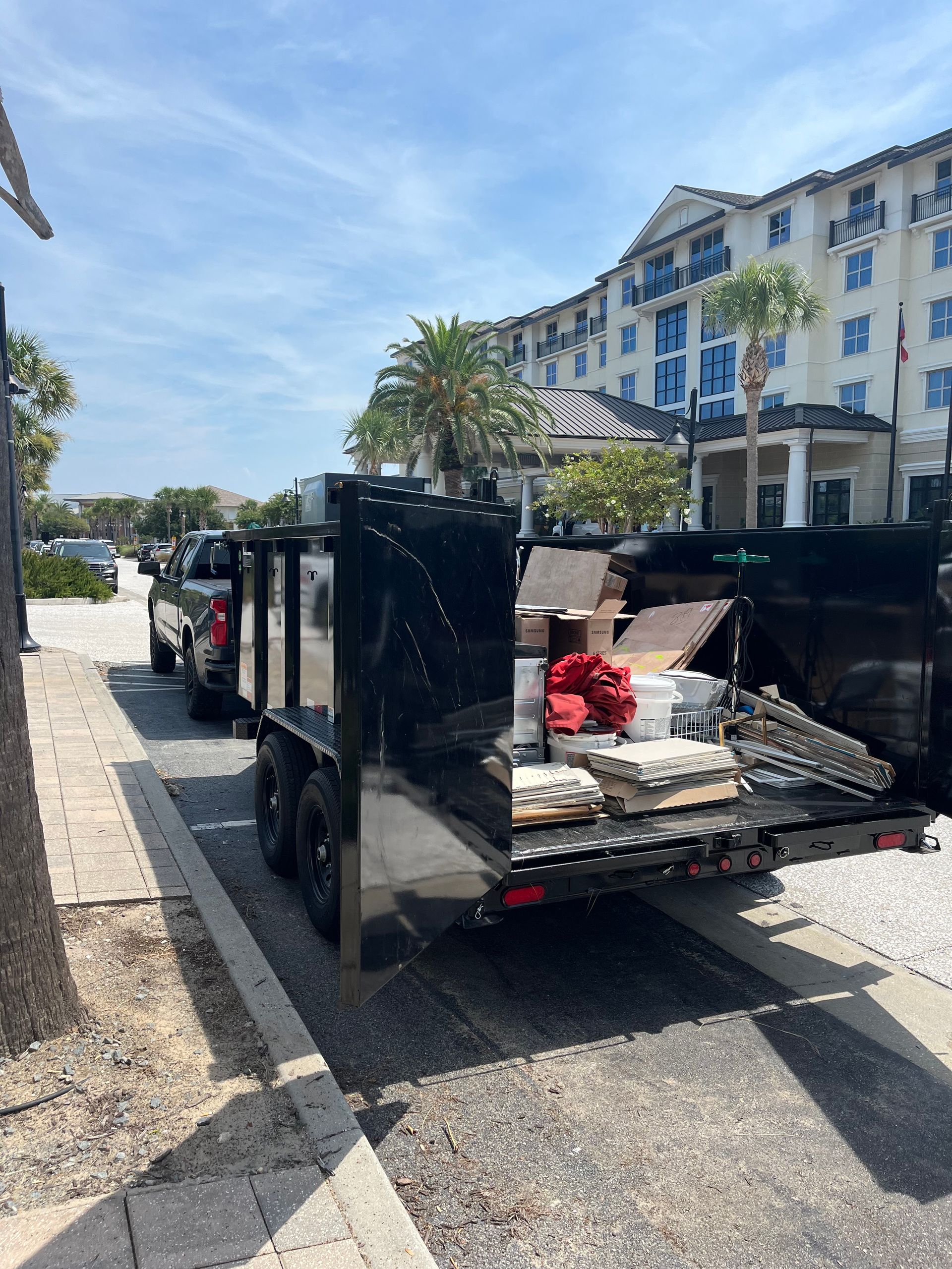 Black truck pulling a loaded trailer parked on a street. Building and palm tree visible in the background.