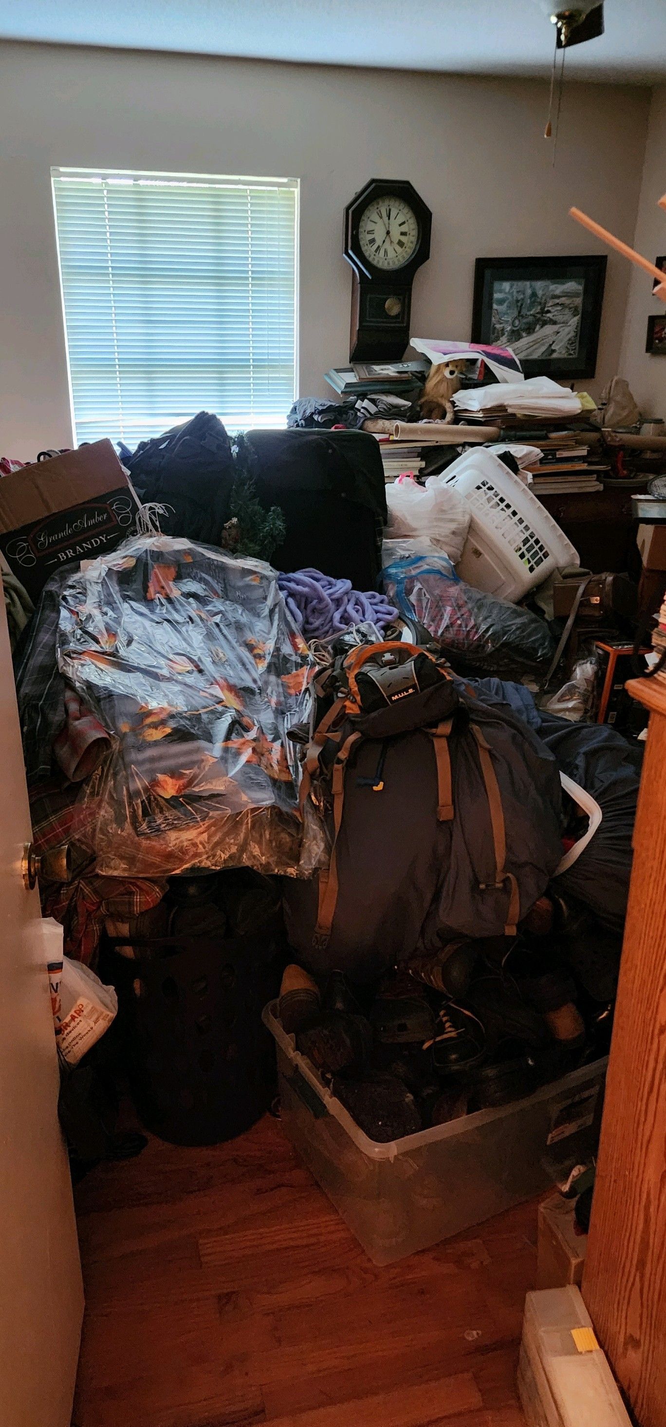 Room filled with clutter. Boxes, clothes, and various items piled up on the floor near a window and a clock.