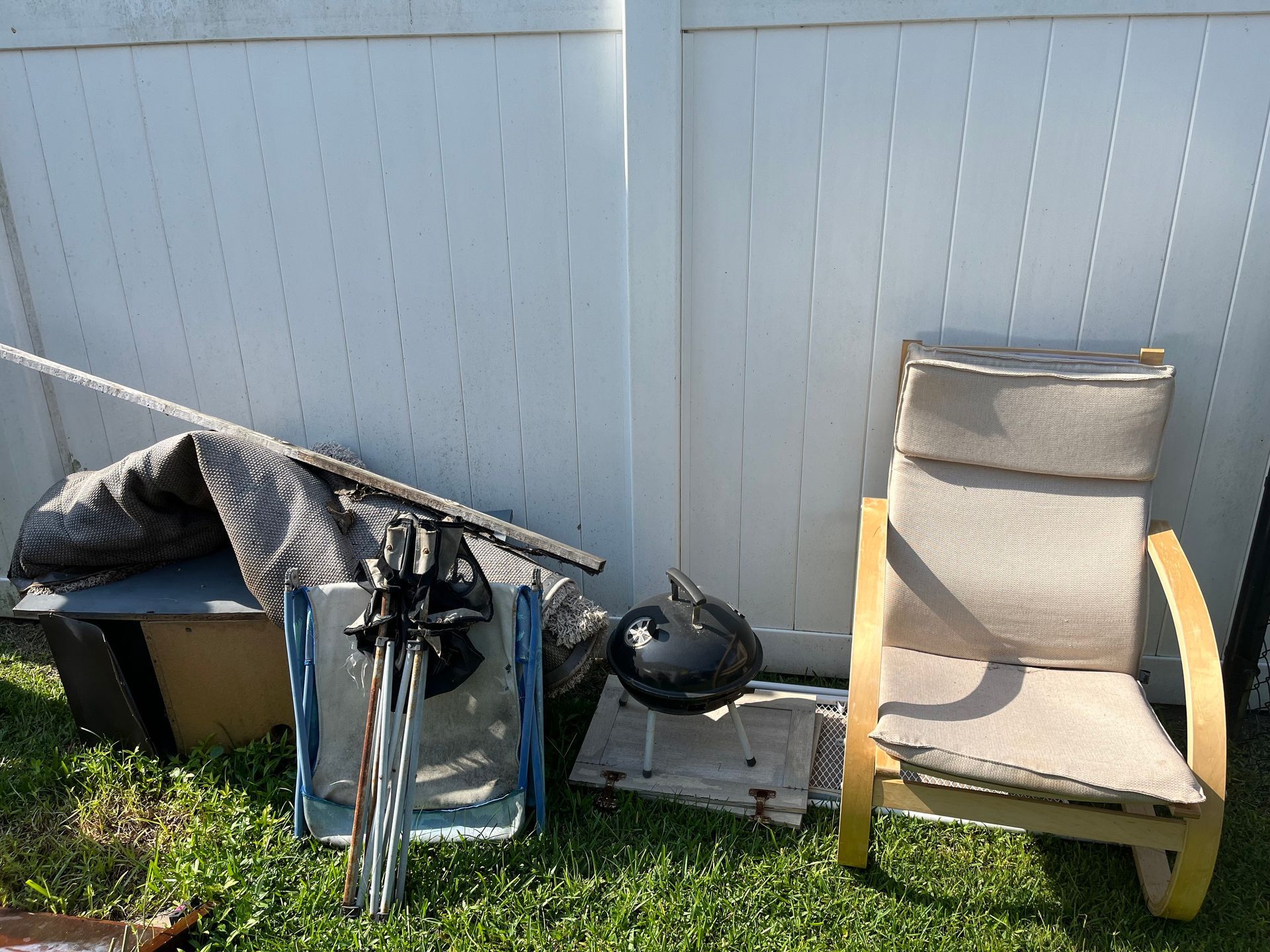 Lawn chair, discarded objects against a white fence on grass in daylight.