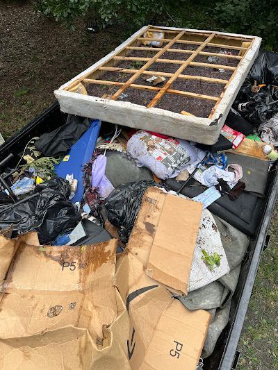 A dumpster overflowing with trash, including a damaged mattress, cardboard boxes, and clothing.