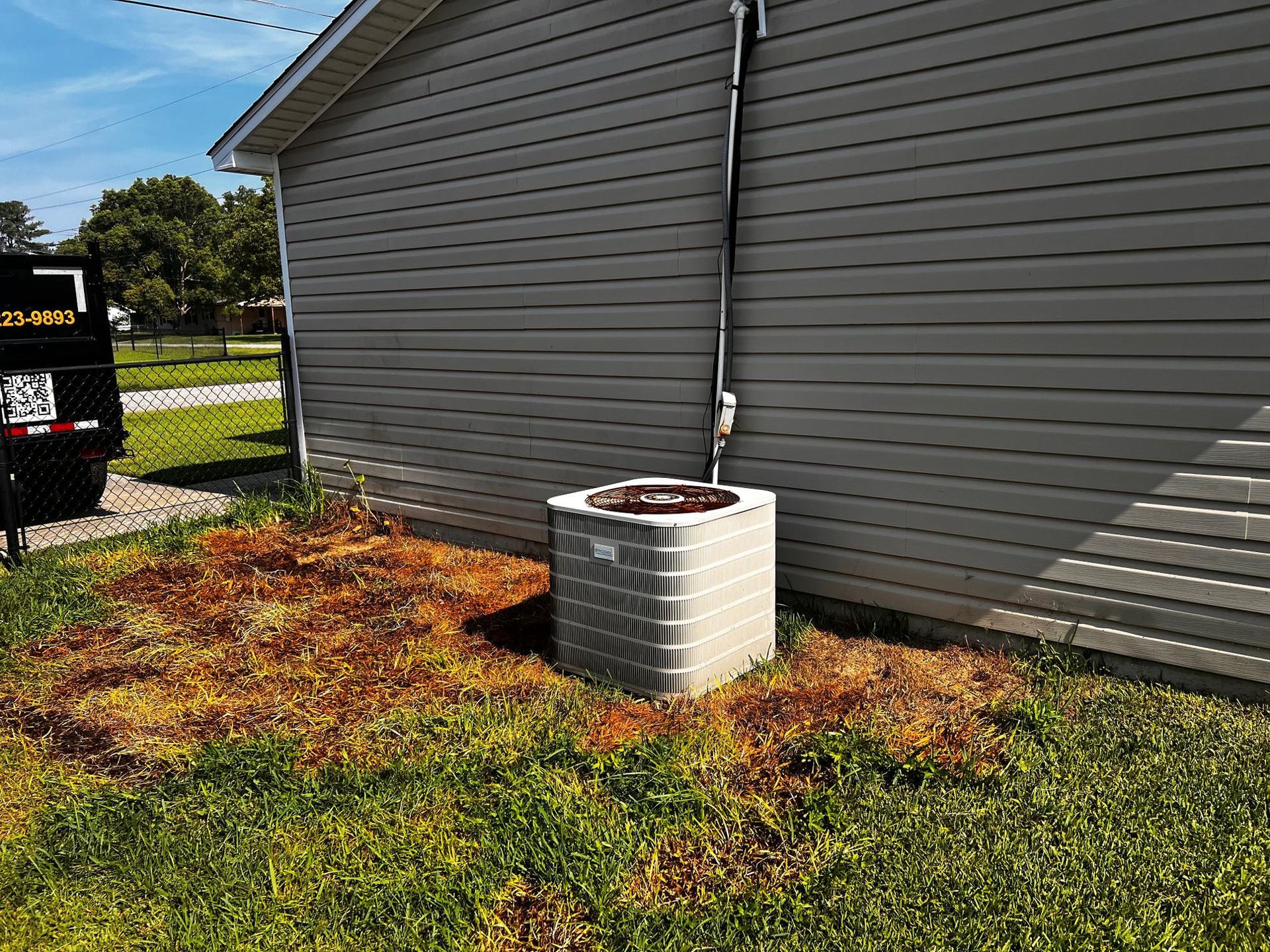 An air conditioning unit sits next to a gray-sided building, with dead grass around it.