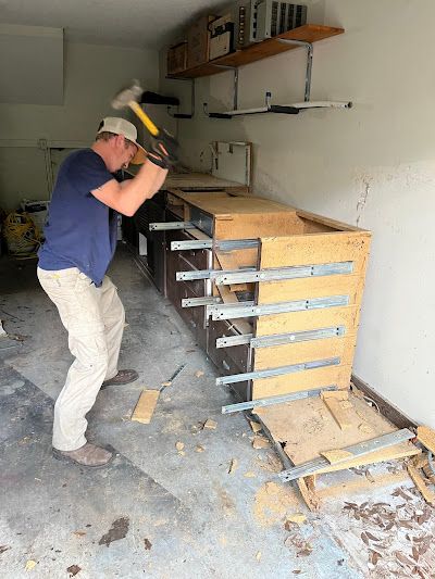 Man in garage, swinging a hammer to demolish a wooden cabinet.
