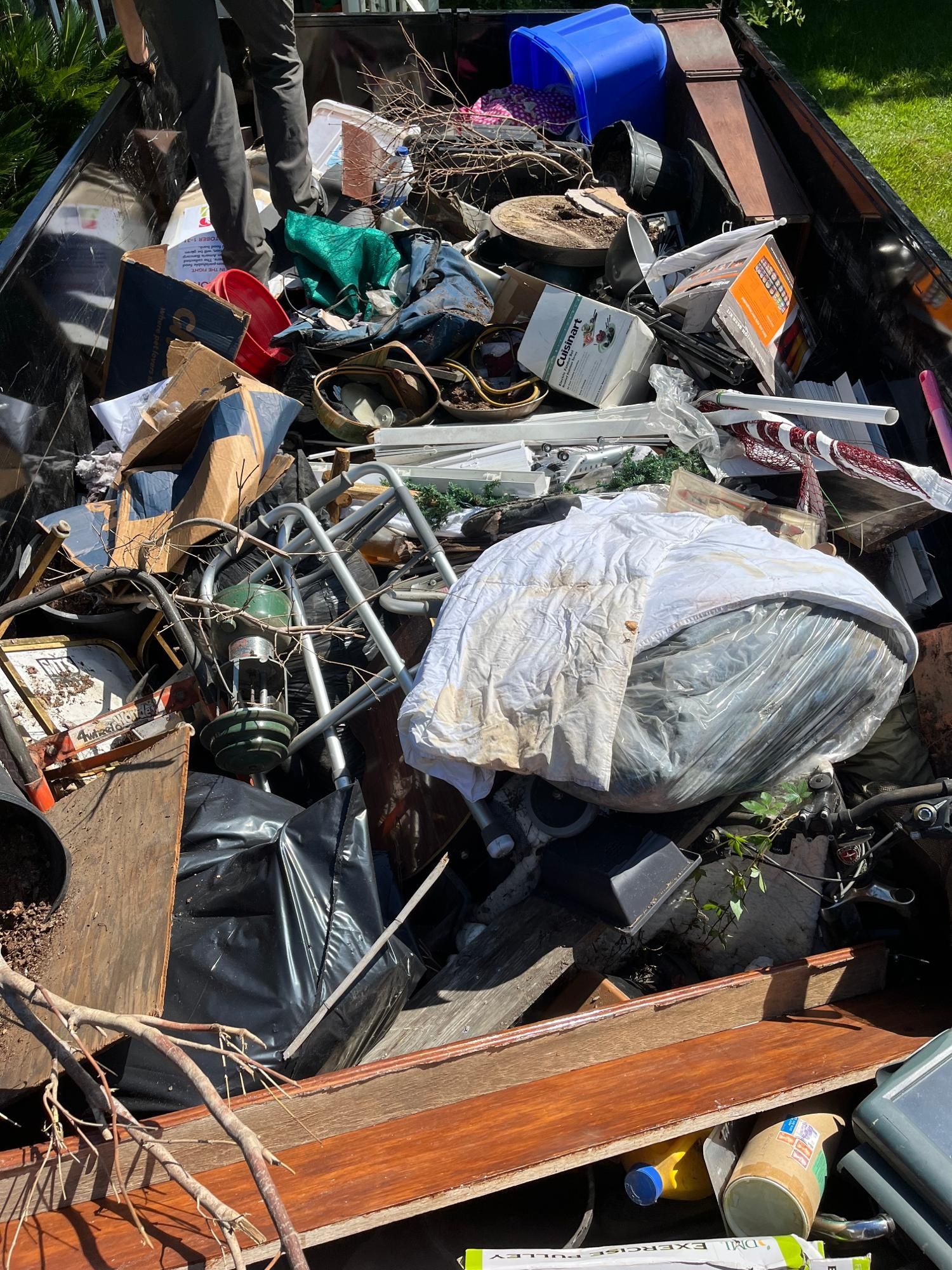 A full dumpster overflowing with trash, including furniture, cardboard, and plastic containers.