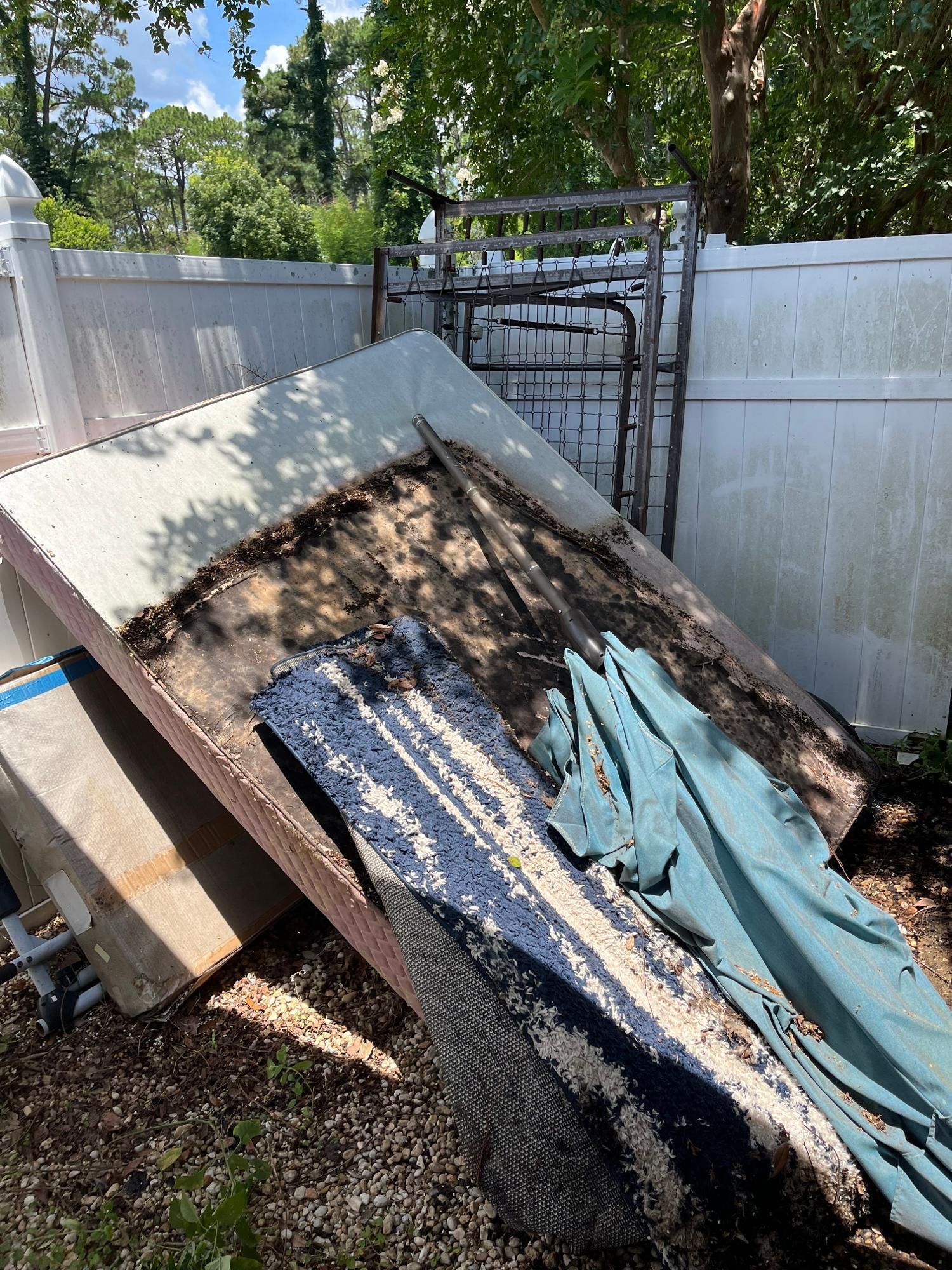 Pile of debris including a metal frame, boards, and cloth against a white fence in a backyard.