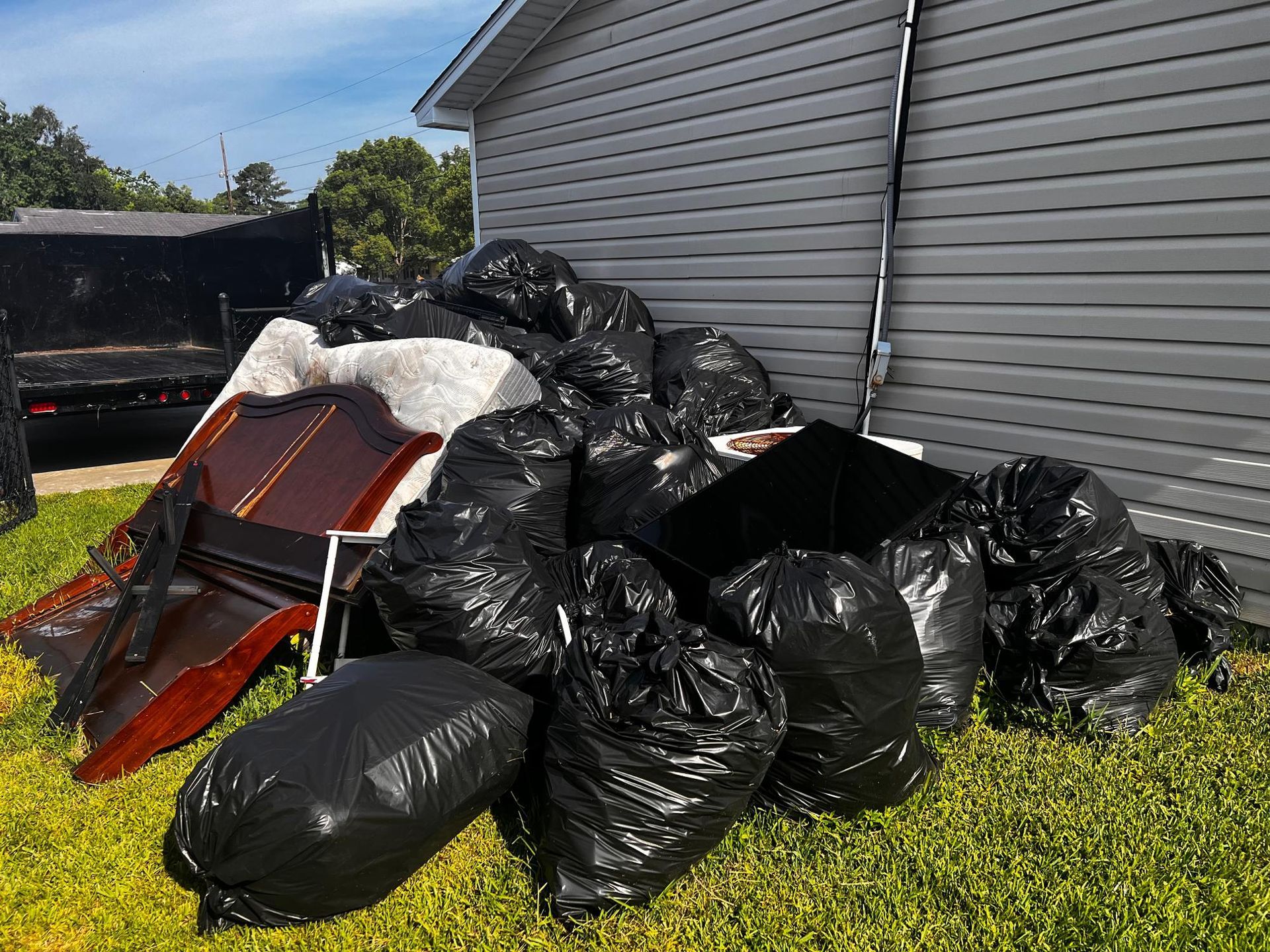 Pile of black trash bags and furniture next to a gray house on green grass.