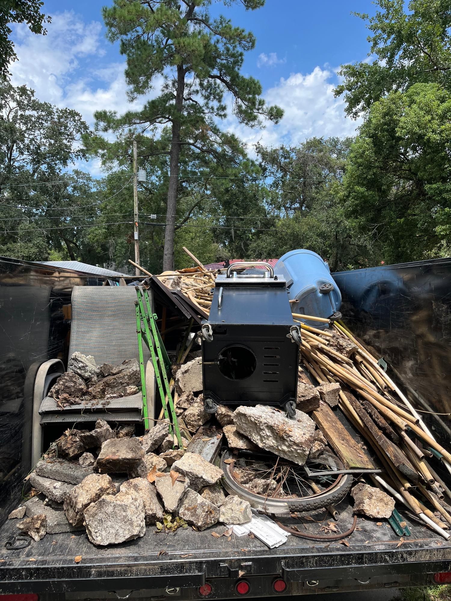 A truck bed filled with construction debris, including rocks, wood, and a black stove, under a blue sky.