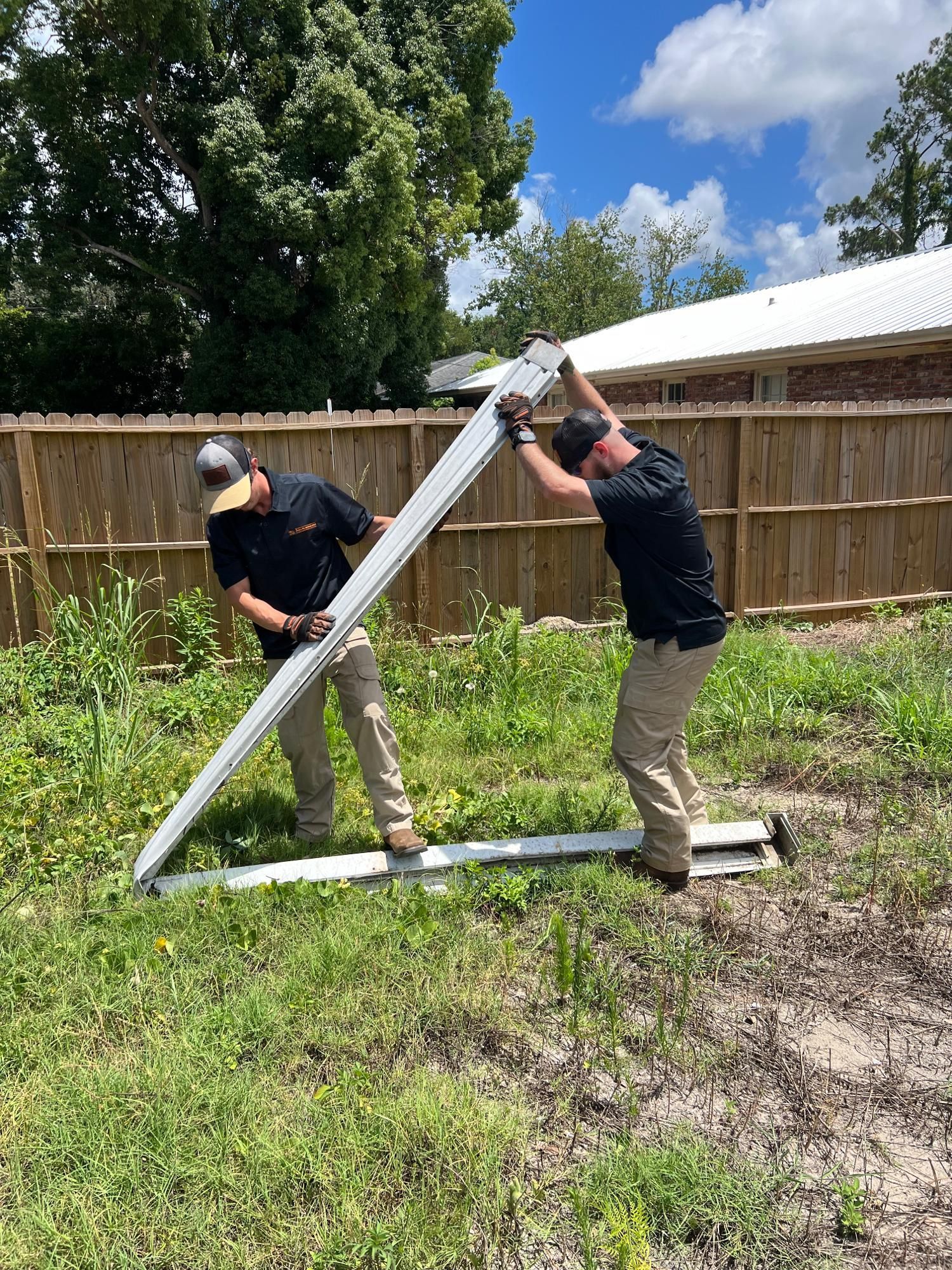 Two men in tan pants and black shirts lifting a long white structure in a grassy area, near a wooden fence.