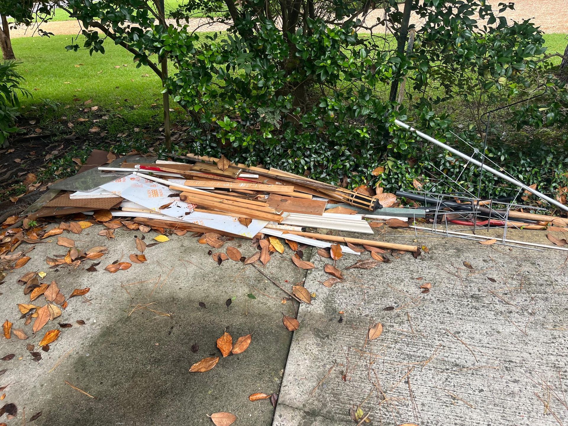 Pile of construction debris on concrete, next to bushes and grass. Fallen leaves scattered around.