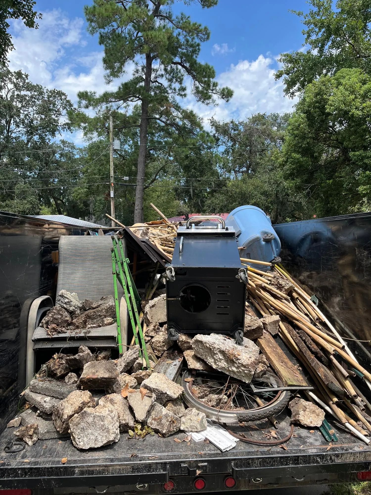 A truck bed filled with debris, including a metal stove, concrete, wood, and a blue water tank, against a backdrop of trees.