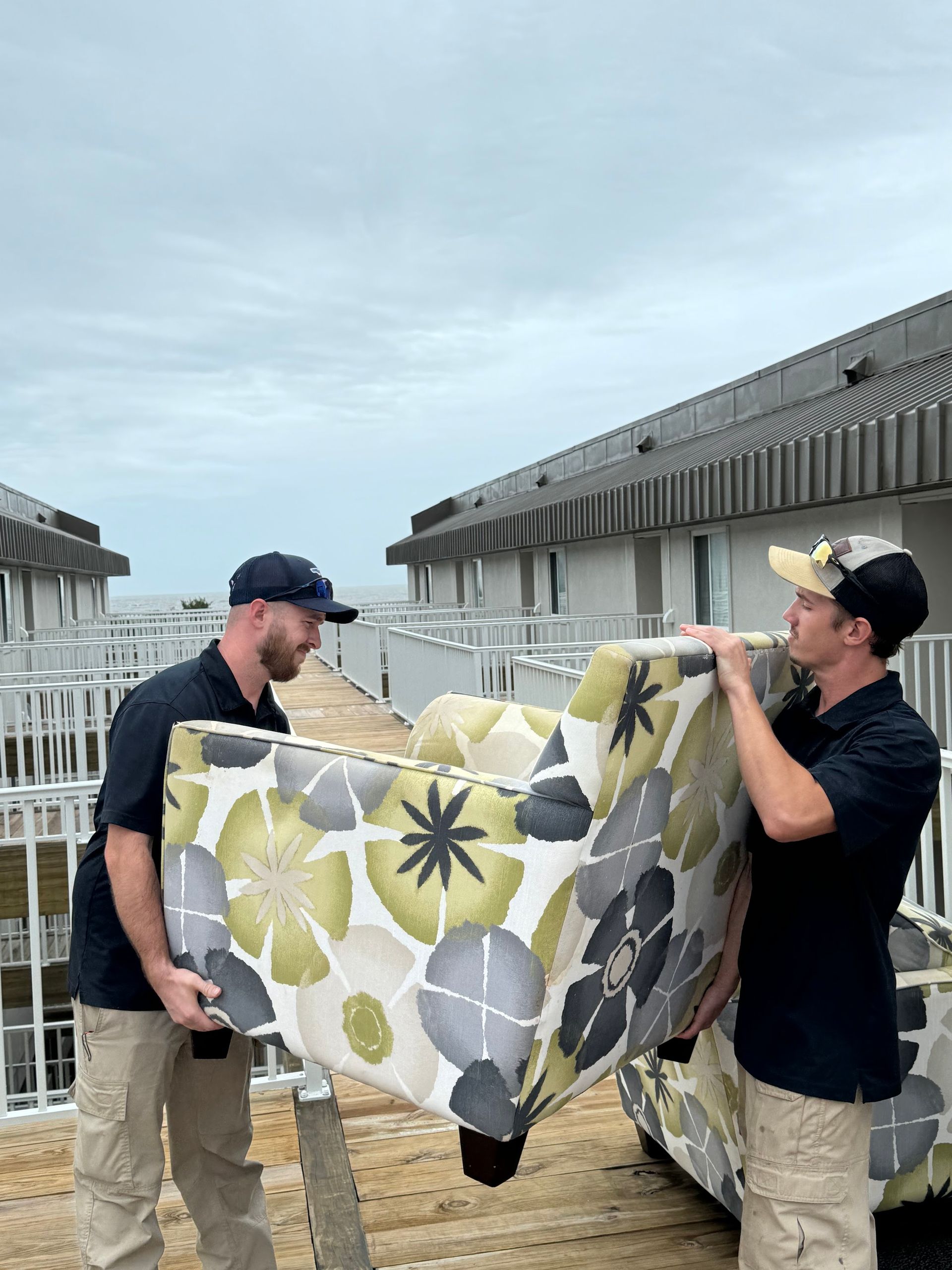 Two men carrying a large floral-patterned couch on a rooftop deck. Cloudy sky and buildings in the background.