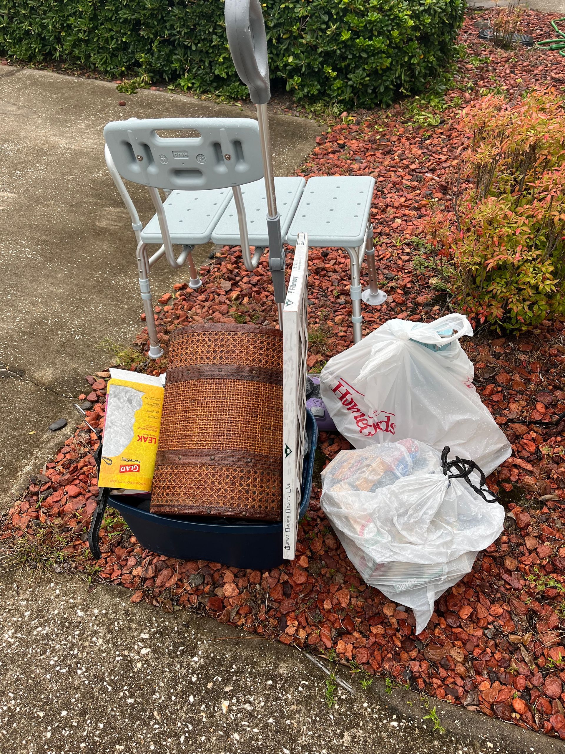 A shower chair, food, and bags of trash sit on a red-mulched walkway next to a bush.