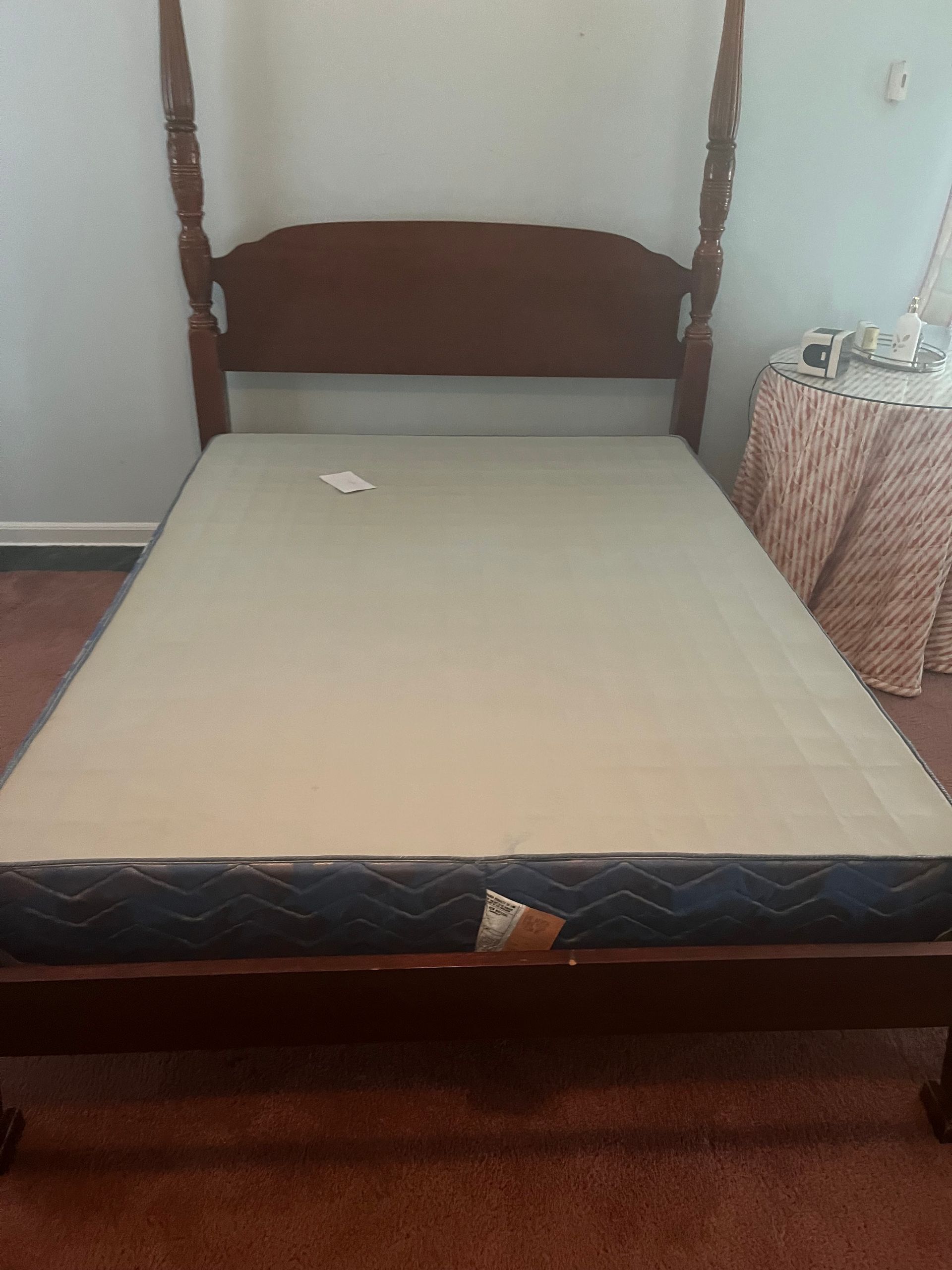 Wooden four-poster bed with beige mattress, set against a white wall, beside a pink table.