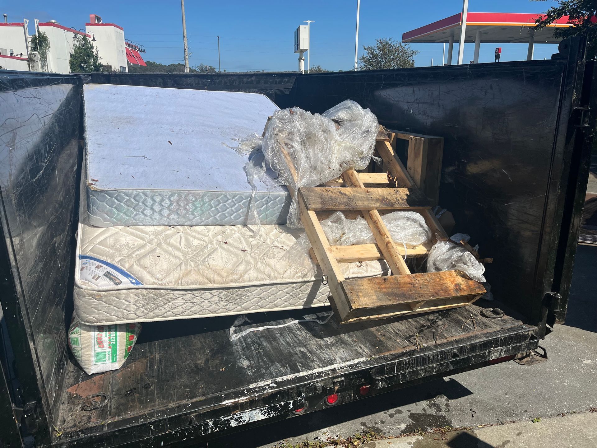 Dumpster filled with discarded mattresses, a pallet, and trash bags, likely near a street.