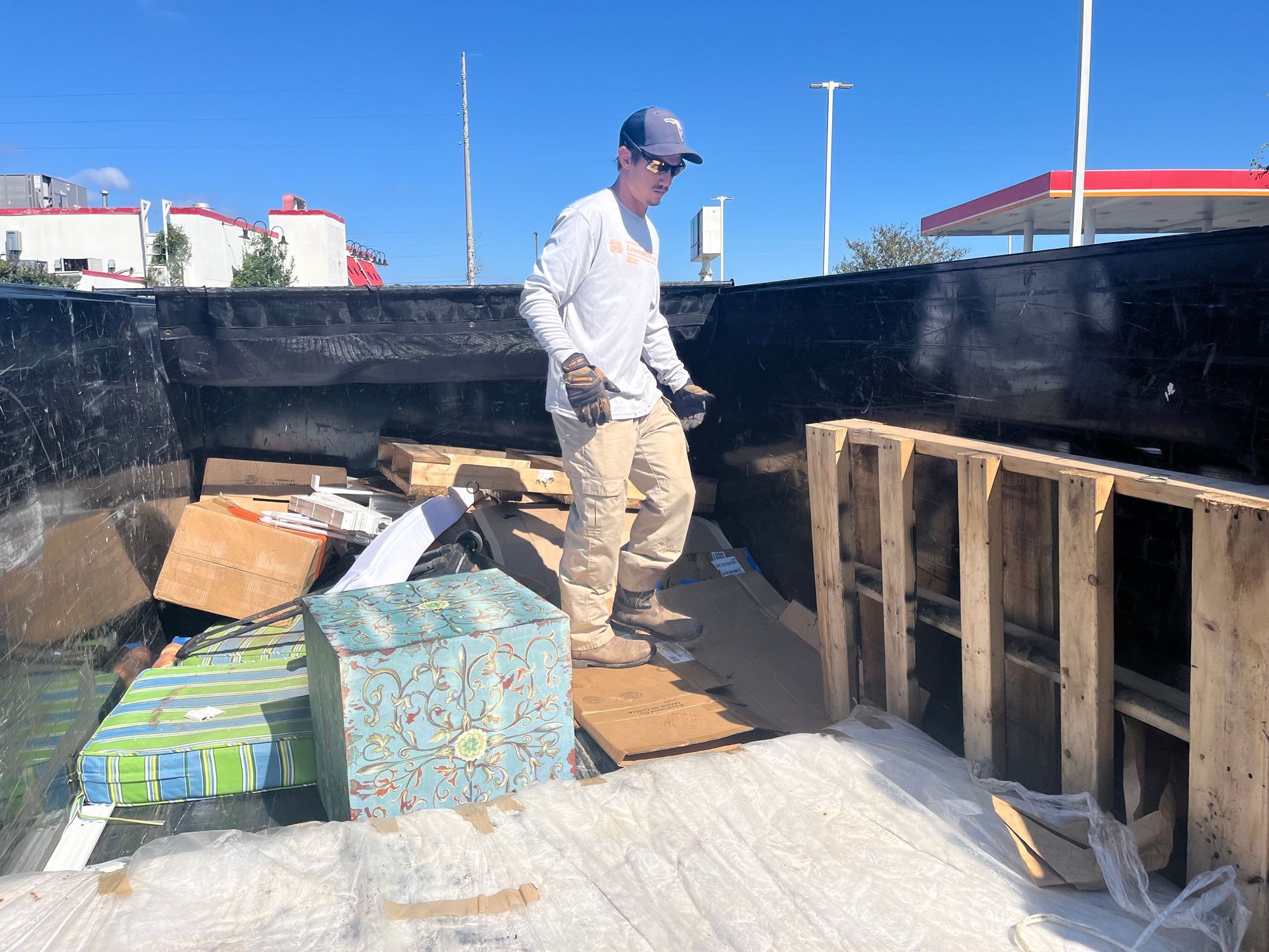 A man stands in a dumpster sorting trash, with pallets and boxes. Bright sunny day.