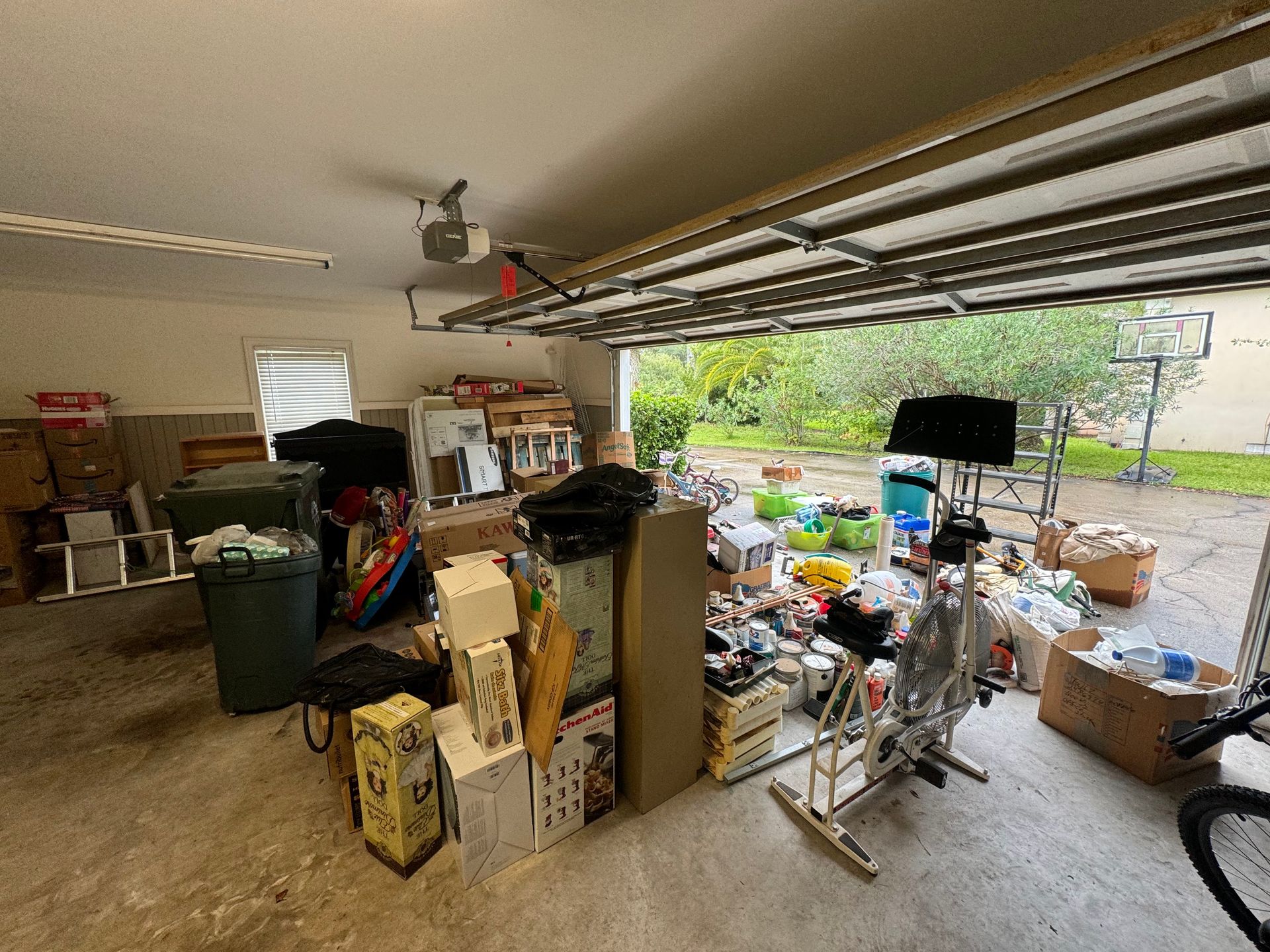 Cluttered garage with boxes, equipment, and open door to backyard with basketball hoop.