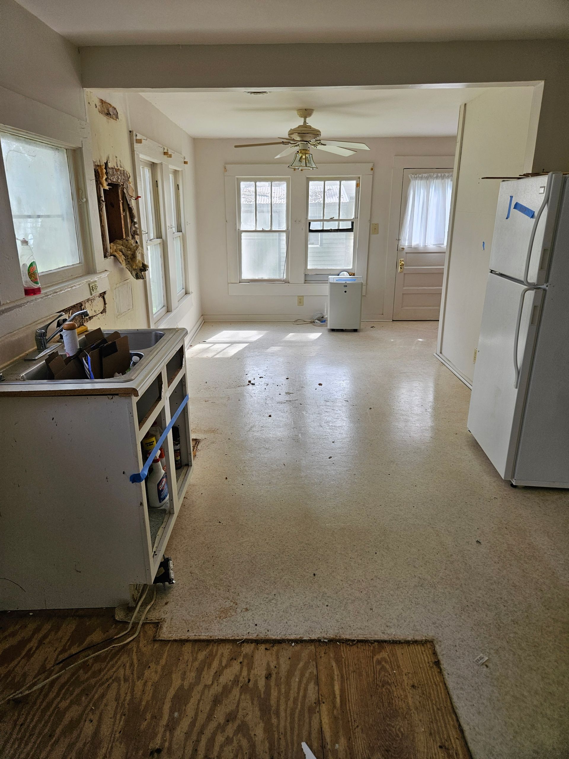 Dilapidated kitchen and living area. Visible damage to walls, cabinets, and flooring.
