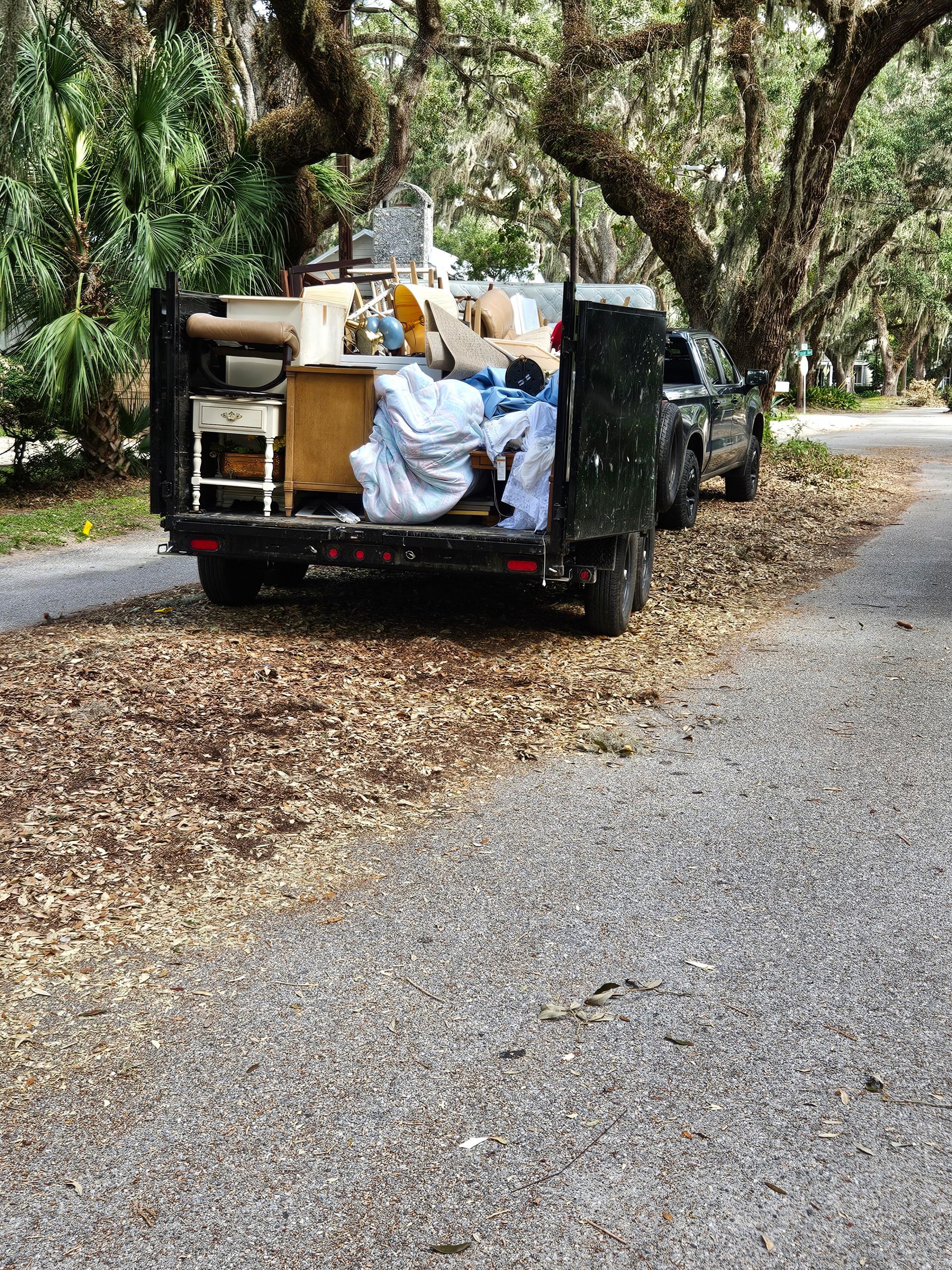 Truck filled with discarded furniture parked on a leaf-strewn gravel driveway under a canopy of trees.