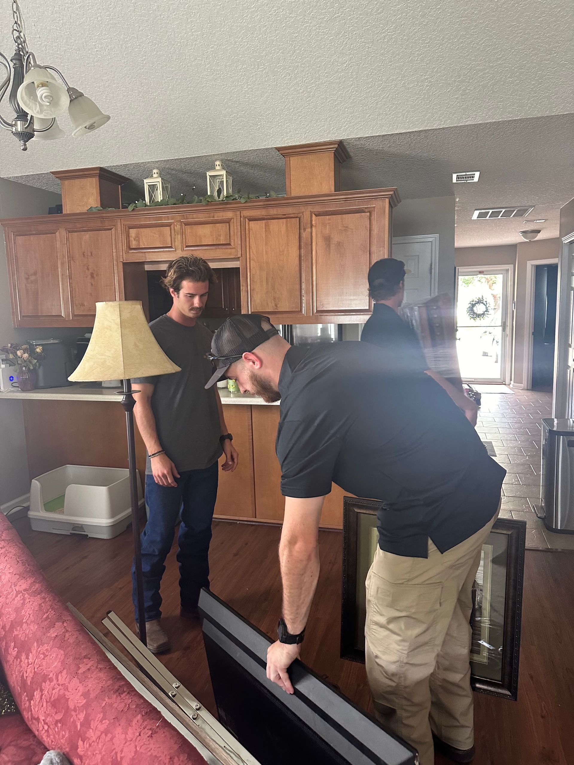 Three people in a house; one holding a TV, others observing near kitchen cabinets.